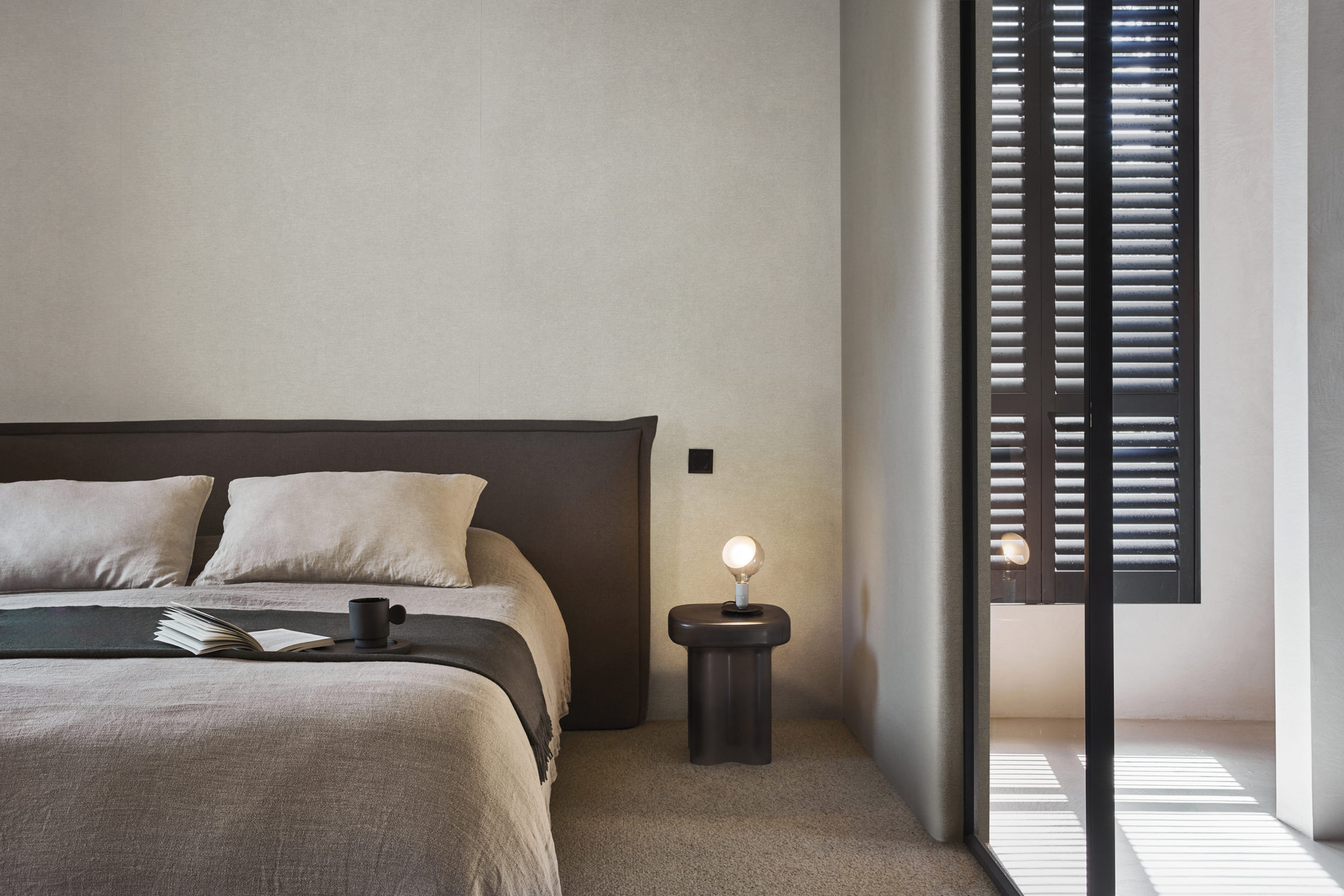 A tranquil bedroom in soothing shades of beige, featuring a glass sliding door separating the sleeping area from the bathroom. A plush, solid light brown carpet enhances the serene atmosphere. The double bed is adorned with an open book and a mug, inviting relaxation. The walls are adorned with Omexco's Silence wallcoverings in a calming beige hue, not only adding aesthetic appeal but also offering acoustic properties for a peaceful and quiet retreat