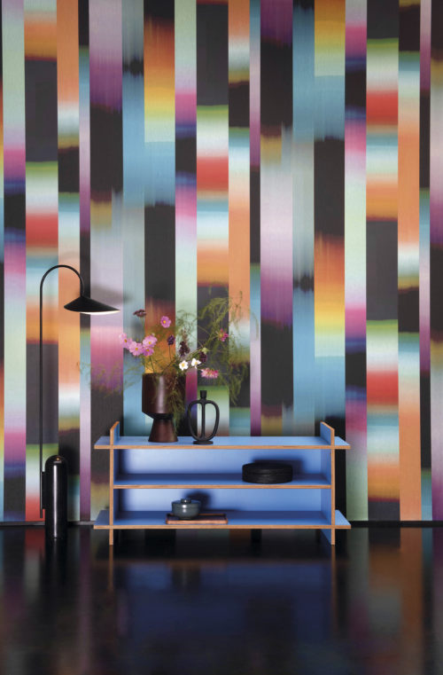 Funky seventies inspired home decor. Bring the unique vibe from an incredible period in time back into your home with the "Time-lapse" wallcovering design from Omexco's "Atelier" collection. This modern rendition of an Ikat weaving show dynamic stripes in a color gradient of orange, turquoise, pink, purple, black and red. The perfect backdrop for an electric blue open cupboard with shelves and minimal black floor lamp.