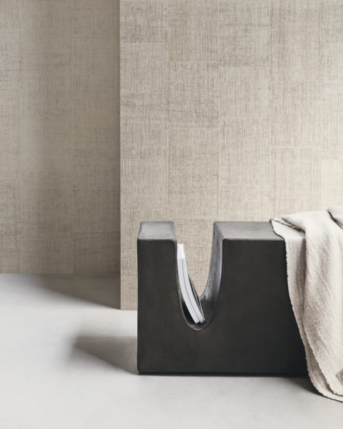 Captivating decor shot featuring Omexco Intuition wallcoverings, meticulously crafted from burlap with an elegant patchwork design and exquisite stitching in an off-white color. The scene is grounded by a gray cast floor, adorned with a wooden minimalist black bench hosting books, topped with a beige blanket gracefully draped over the seat.
