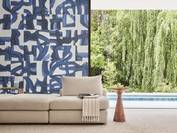 A stunning living room featuring Omexco's Rivage wallcovering in a captivating blue hue with artistic brushstrokes. The design skillfully weaves echoes of ropes and maritime knots, paying tribute to the intricacies of seafaring. In the foreground, a close-up of a beige sofa and a minimalist wooden side table exudes modern elegance. Beyond, an open window reveals a refreshing view of a swimming pool, adding to the overall sense of tranquility and sophistication in this beautifully adorned space.