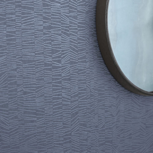 A captivating close-up of a wall adorned with Omexco's high-performance wallcovering collection Rivage in a soothing blue hue. The design boasts textured tonal stripes that beckon with subtle elegance. A round mirror hangs gracefully on the wall, reflecting the sophisticated ambiance. These PVC-free wallcoverings not only offer bleach cleanability but also feature enhanced performance with scrubbability, ensuring both style and durability.