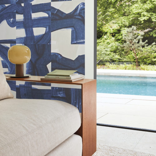 A stunning living room featuring Omexco's Rivage wallcovering in a captivating blue hue with artistic brushstrokes. The design skillfully weaves echoes of ropes and maritime knots, paying tribute to the intricacies of seafaring. In the foreground, a close-up of a beige sofa and a minimalist wooden side table exudes modern elegance. Beyond, an open window reveals a refreshing view of a swimming pool, adding to the overall sense of tranquility and sophistication in this beautifully adorned space.