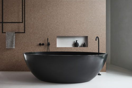 A luxurious bathroom featuring a striking black freestanding oval-shaped bathtub with modern taps and accessories. The walls are adorned with Omexco's high-performance wallcovering collection, Rivage, boasting textured tonal stripes in a captivating brown/red hue. The design adds a touch of sophistication and warmth to the space. Behind the bathtub, a niche in the wall provides a stylish storage solution.