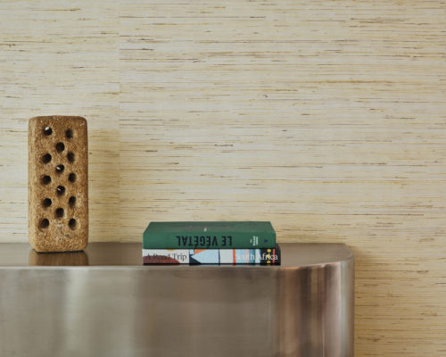 An elegant decor composition featuring a silvery metal cupboard positioned against a neutral wall adorned with Omexco's handmade natural Intuition wallcovering in a soothing beige ramie weave. On top of the cupboard, two carefully placed books add a touch of sophistication, complemented by a brick-like decorative element, creating a harmonious blend of textures and tones.