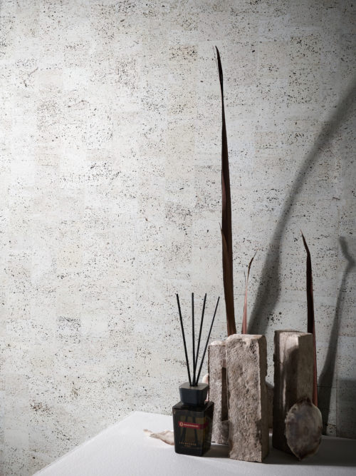 An image of light grey cork Omexco wallcoverings with a white surface is displayed. In the foreground, there are various decorative elements, including a bottle of house perfume, three upright bricks and some shells. The wallcovering has a textured surface with a natural cork pattern, and the light grey color gives it a modern and elegant look. It belongs to the “Casca” collection, a curated selection of the most stunning variations of mulberry and cork oak barks.