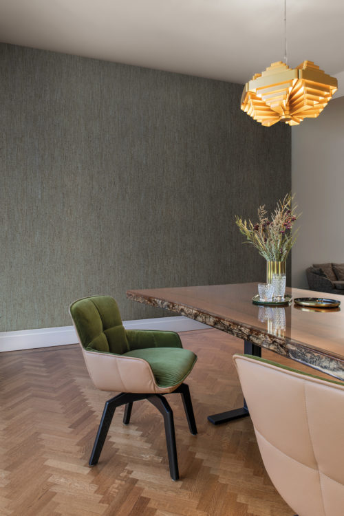An image of a beautifully designed dining room featuring a herringbone patterned wooden floor. A gold hanging lamp illuminates the space, casting a warm and inviting glow. The centerpiece of the room is a wooden dining table adorned with a lovely bouquet of dried flowers. Surrounding the table are two comfortable dining chairs in beige and green. The walls are adorned with a textured wallpaper, adding a touch of sophistication to the space. These wallcoverings belong to the exquisite collection “Casca” by Omexco, exposing the soft and matt natural textures of cork. The raw material is enhanced in a graphic composition of grey and green hues.