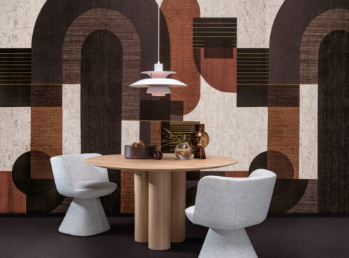Example of a dining room with a bold graphic wallcovering showing a pattern of geometric shapes in grey, black, burnt orange and white hues. This panoramic design is digitally printed on cork and belongs to Omexco’s wallcovering collection “Casca”. We see a round wooden dining table in the center of the room with a white pendant lamp hanging above it. Decorative elements such as small glass vases and a bowl filled with walnuts are placed on the tabletop. Two soft grey dining chairs are positioned around the table. The overall aesthetic is modern with a touch of warmth and texture.