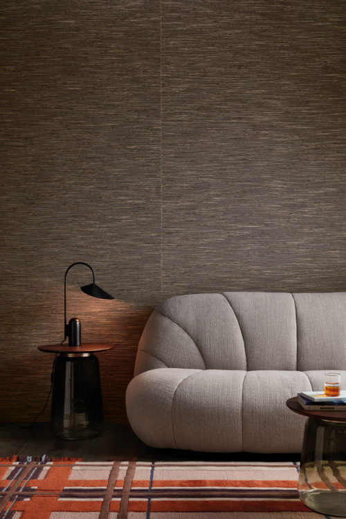 An image of an inviting living room with a soft grey couch as the focal point, flanked by two round glass sidetables, one topped with a black table lamp. A colorful carpet in shades of orange, pink and brown lies on the floor, adding a pop of color to the room. The walls are covered in textured dark grey Omexco wallcoverings, providing a stylish backdrop for the cozy seating area. The wallcovering consists of wool and linen with a rich textural quality and belongs to the textile collection “Loom Stories".