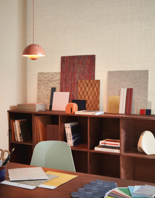 Interior designer's or architect’s inviting home office with a massive wooden desk and shelving unit, pink vintage Flowerpot VP1 Hanging Lamp and baby blue Eames DSW plastic chair. We see samples of Omexco wallcoverings and other materials, books and small storage boxes on the desk and in the shelving unit. On the wall, Omexco’s textile wallcovering collection “Loom Stories” is installed: a very textured weave of thick, off white yarns. A soothing neutral wall you would love to touch.