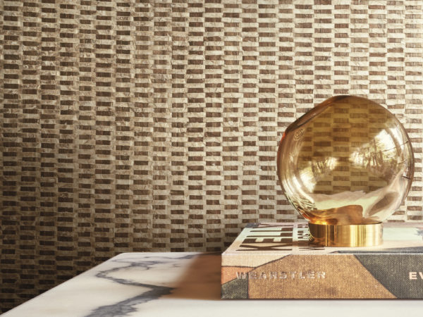 An elegant round glass table lamp, beautifully positioned on top of a book, serves as the focal point in this stylish decor. The backdrop features luxurious capiz wallcoverings from Omexco's Archipelago collection, showcasing a captivating blend of gold and brown shades, creating a sophisticated and inviting atmosphere.
