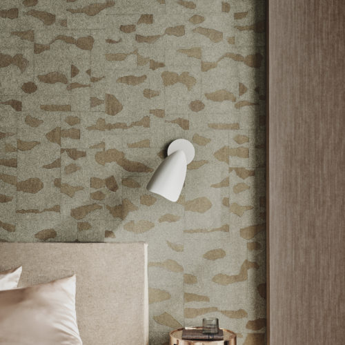 This close-up of a chic bedroom showcases an inviting bed adorned with champagne-colored sheets, accompanied by a luxurious gold metal nightstand and a sleek white wall lamp. The backdrop features fine stardust Omexco wallcoverings, where mica is delicately printed with an organic design, evoking a sense of nature. The combination of soft hues, metallic accents, and nature-inspired textures creates a serene and sophisticated atmosphere in this stylish bedroom setting.