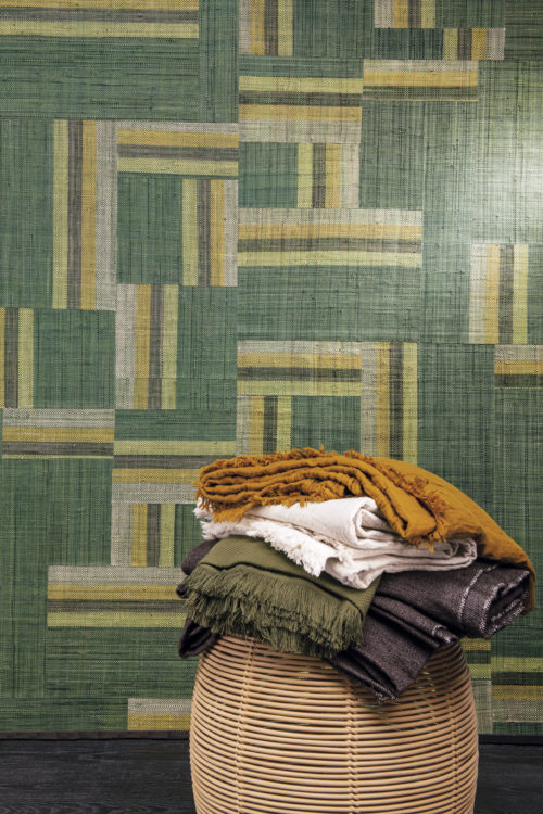 Authentic craftmanship embodies the bold Atelier collection designed by the creative minds of Omexco. This image shows an impressive green patchwork of handwoven raffia. In front of the wall we see a ratten basket with a stack of folded blankets in shades of ochre, khaki, beige and grey.