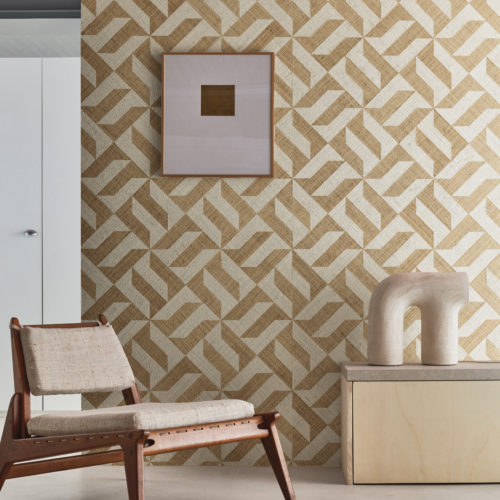 A captivating decor shot featuring an ethnic Scandinavian-style wooden chair on the left and a light wood cupboard on the right adorned with a ceramic decorative element. The grey cast floor provides a neutral foundation, while the backdrop showcases Omexco's Intuition wallcoverings—a raffia patchwork displaying a striking geometric design with yellow and white shapes, creating a harmonious and stylish interior composition.