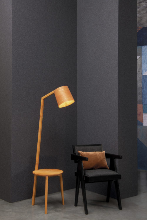 Contemporary interior with a black chair with orange pillow, wooden end table with table lamp and a grey concrete floor. The wall shows a dark grey plain mica wallcovering by Omexco.