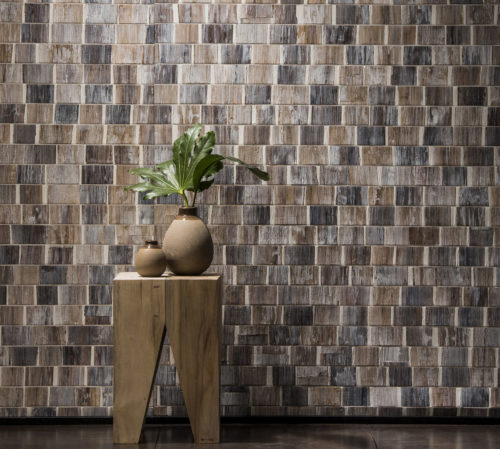 Detail of a white wash bakbak wallcovering. In front of it a wooden stool with triangle opening. Two nude vases with green leaves.
