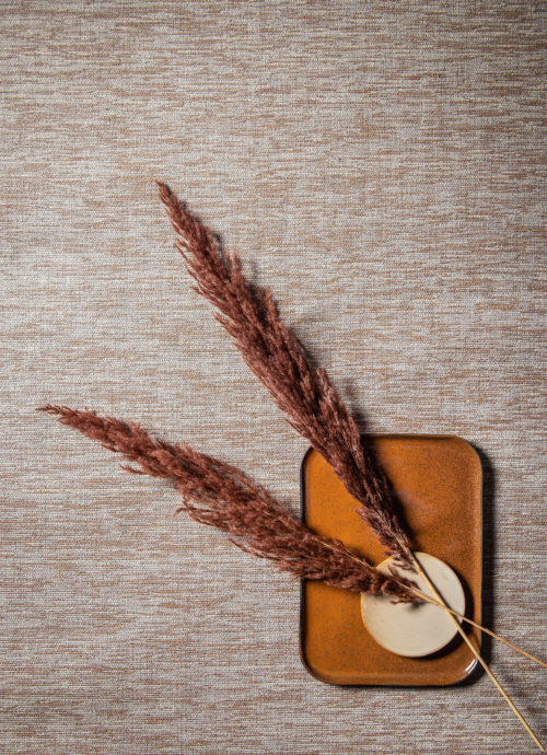 Paper yarn delicately woven into seamless wallcoverings in sand tones. We see a closeup of the wallcoverings as well as decorative feathers and ceramic plates in different shapes.