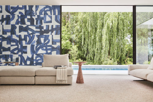 A stunning living room featuring Omexco's Rivage wallcovering in a captivating blue hue with artistic brushstrokes. The design skillfully weaves echoes of ropes and maritime knots, paying tribute to the intricacies of seafaring. In the foreground, a close-up of a beige sofa and a minimalist wooden side table exudes modern elegance. Beyond, an open window reveals a refreshing view of a swimming pool, adding to the overall sense of tranquility and sophistication in this beautifully adorned space.