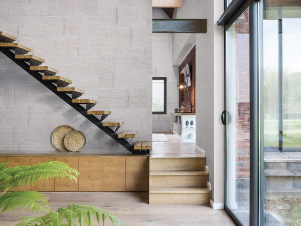 Wall with raffia blocks wallcovering. Stairs run diagonally through the picture. Large window on the right. We get a look at the tv room in the back with brick wall and bird statues.