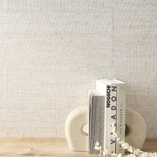 This close-up highlights the refined elegance of Omexco's Pure collection, reference PU211, known as Anima. The wallpaper features a raffia weave with an irregular texture, beautifully enhanced by a delicate layer of white paint. The natural imperfections in the weave add depth and character, creating a timeless aesthetic. The wooden surface in the foreground holds a sculptural bookend and an assortment of art and design books, complemented by delicate white florals. Together, the textured wallcovering and curated decor exude understated sophistication, perfect for contemporary interiors.