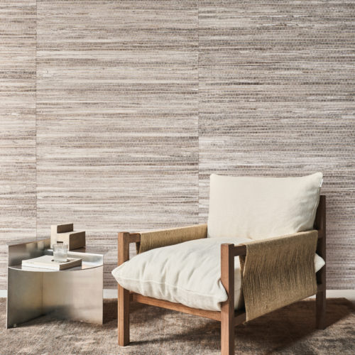 This serene reading corner features Omexco's Pure collection, reference PU222, known as Innate. The wallcovering showcases a refined abaca weave, skillfully crafted in a choice of rich, thick texture or a finer, more delicate variant, both enhanced with a subtle coat of white paint. The natural striations create depth and add a warm, organic feel to the space. A minimalist wooden armchair with cream cushions is paired with a silver metallic side table, holding a stack of books and a glass. The neutral tones of the wall and furnishings complement each other, creating an inviting yet understated aesthetic.