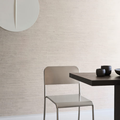 A minimalist dining area featuring Omexco's Pure collection, reference PU231, known as Osmos. This refined wallpaper showcases a delicate weave of natural waterlily fibres, gracefully finished with a soft layer of white paint. The understated design brings a sense of calm and sophistication to the space. A sleek metallic chair pairs harmoniously with a modern dark-stained dining table, complemented by minimalist ceramic bowls in muted tones. On the wall, a sculptural white art piece adds an abstract element, balancing the natural textures of the wallpaper. This setting seamlessly blends contemporary aesthetics with the organic beauty of natural materials.
