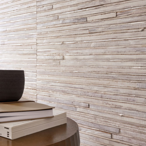 A modern close-up interior composition featuring Omexco's Pure collection, reference PU241, named Native. This striking wallpaper showcases a bold weave of wide abaca strips, meticulously coated with white paint, creating a textured, layered effect that exudes natural sophistication. The scene includes a minimalist table adorned with a dark ceramic bowl, stacked books, and a leather notebook, offering a harmonious balance of natural and contemporary elements. The wallpaper's tactile finish adds depth and character to the space, embodying a refined yet raw aesthetic that complements minimalist and earthy interiors.