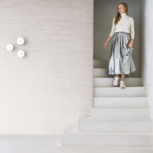 A minimalist and contemporary interior showcasing Omexco's Pure collection, reference PU251, named Delicate. This refined wallpaper features fine raffia elevated by subtle metallic threads, blending natural texture with a hint of shimmer for a sophisticated aesthetic. A woman descends a white staircase wearing a cream turtleneck sweater and a striking silver metallic skirt, adding a touch of modern elegance to the serene space. The backdrop enhances the tranquil setting with its understated yet intricate texture, making it ideal for minimalist interiors that value subtle luxury and artisanal craftsmanship.
