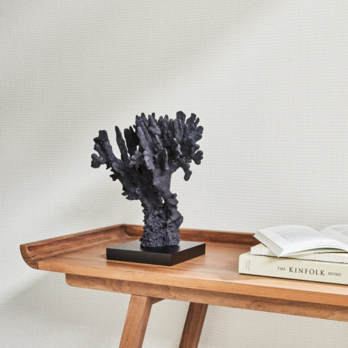 A close-up view of a wall adorned with Omexco's high-performance wallcovering collection, Rivage, showcasing a subtle plain white design from the collection. Against the wall, a Scandinavian-style wooden side table holds three books as well as a captivating coral sculpture. The PVC-free Rivage wallcoverings not only offer bleach cleanability but also feature enhanced performance with scrubbability, ensuring both style and durability.