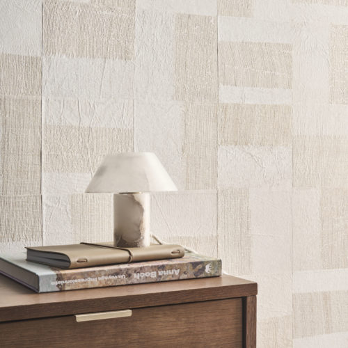 A refined interior featuring Omexco's Pure collection, reference PU261, a wallcovering named Intensify, showcasing a geometric paper patchwork enriched with linen-toned openwork abaca weaving. This design adds depth, texture, and a tactile dimension to the space, creating a striking visual statement. The scene includes a minimalist wooden cabinet with a subtle brass handle, topped with a stack of books and a leather notebook, reflecting thoughtful organization. A marble table lamp with a sculptural quality enhances the luxurious yet understated aesthetic. The harmonious combination of organic textures and geometric patterns makes this wallpaper a focal point for modern, sophisticated interiors.