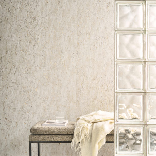 An elegant and serene space featuring Omexco's Pure collection, reference PU271, a wallcovering named Nebbiosa that highlights the subtle plain texture and the unique grain of cork with shimmering delicacy. The natural tones of the wallpaper add a soft, organic ambiance to the room, enhanced by the reflective glass block partition that introduces additional light and texture. The scene includes a modern bench with a textured fabric seat, adorned with a cream-colored throw and a neatly placed book with a glass tumbler on top. This harmonious composition celebrates natural materials and understated elegance, ideal for contemporary and minimalist interiors.
