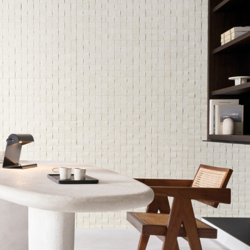 A sophisticated office setting featuring Omexco's Pure collection, reference PU281, a bold wallcovering that showcases a weave of mulberry bark, highlighting the inherent richness and texture of this natural material. The intricate square grid design adds a tactile element and visual depth to the space, emphasizing the organic beauty of the bark. The room includes a sculptural white desk with soft curves, adorned with a modern black desk lamp and a tray holding minimal ceramic cups. To the right, a mid-century inspired wooden chair with cane detailing complements the natural tones of the wallpaper. Built-in shelves displaying books and decor seamlessly integrate with the overall minimalist aesthetic, creating a tranquil and stylish workspace.