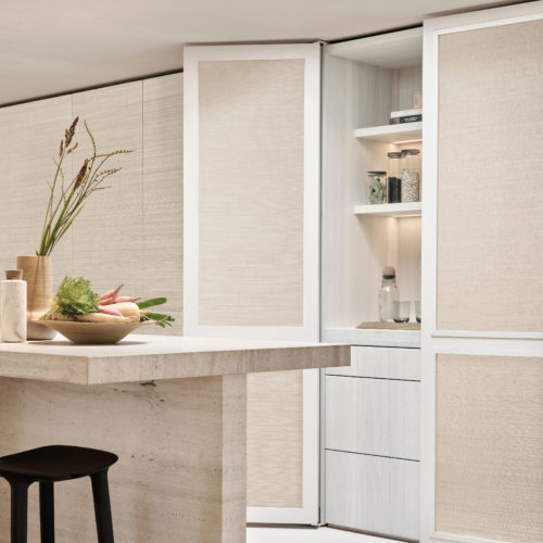 A sophisticated kitchen interior featuring Omexco's Pure collection, reference PU292, a wallcovering showcasing sisal in its most elegant simplicity. The natural texture of the wallpaper adds depth and warmth, creating a serene and refined ambiance. The scene includes a central stone kitchen island with clean, minimalist lines, paired with a sleek black barstool for a touch of contrast. On the countertop, a wooden bowl filled with fresh vegetables and a ceramic vase with dried grasses highlight the harmony between nature and design. In the background, paneled cabinets with integrated shelving and sisal detailing seamlessly blend functionality with aesthetic appeal, offering a perfect balance of practicality and elegance.