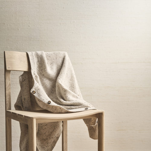 A minimalist and elegant composition showcasing Omexco's Pure collection, reference PU311, a wallpaper crafted from precious silk, revered as the most prized of all natural fibers. The refined texture of the wallcovering exudes understated luxury and adds a natural sophistication to the space. The scene features a wooden chair with clean lines, draped with a textured beige knit cardigan, blending warmth and softness with the raw beauty of the silk wallpaper. The light wooden flooring complements the neutral tones of the wall and furniture, creating a harmonious and serene atmosphere that celebrates natural materials.