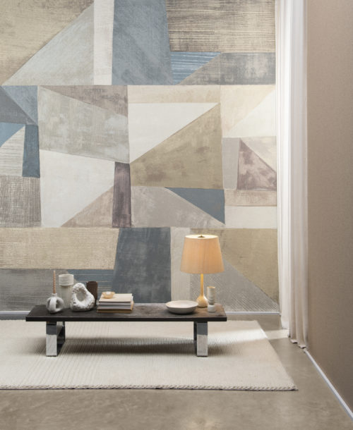 Moonstone by Omexco is a lavish collection of textured wallcoverings softly iridescent like the Moonstone. In this picture we see a feature wall with an XXL abstract art printed on fine mica. The geometric design with diagonal lines is colorful, yet not overwhelming with its soft shades of beige, white, grey, soft pink and unsaturated blue. In front of the wall, we see a low wooden coffee table with small vases and other ceramic decorations as well as a pile of books and a table lamp.