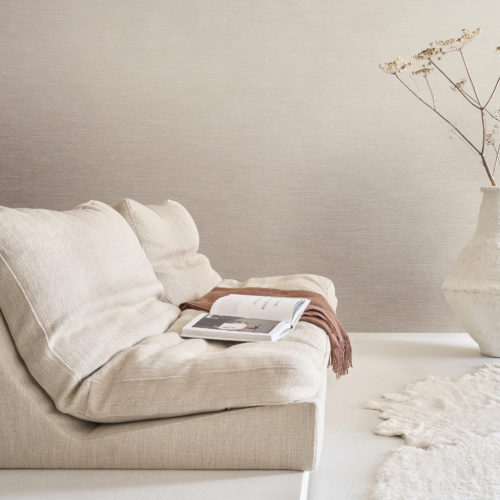 A serene living room featuring Omexco's Pure collection, reference PU351, composed of twisted textile warps crafted from noble materials such as linen. The wallcovering’s refined texture adds subtle depth and sophistication, creating an understated yet elegant atmosphere. The scene includes a low, plush sofa in soft beige tones, styled with a draped rust-colored throw and an open book. To the right, a large, sculptural white vase holds delicate dried flowers, while a soft, irregularly shaped white rug enhances the minimalist aesthetic of the space. The overall design harmoniously combines natural materials and muted tones to evoke a tranquil, refined interior.