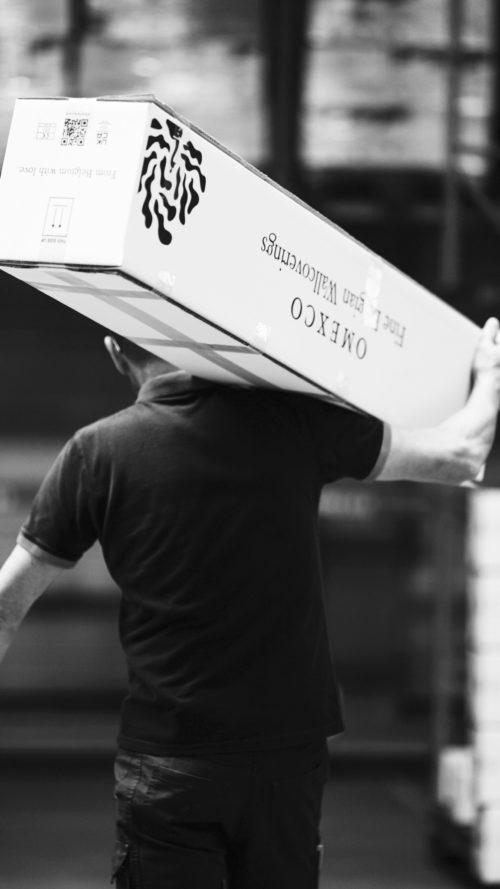 A black-and-white photograph of a warehouse worker carrying a large Omexco box on his shoulder. The packaging, branded with the company's logo and tagline "From Belgium with love," highlights Omexco’s commitment to quality and craftsmanship. The blurred background suggests a dynamic logistics environment, ensuring that every order is handled with care.