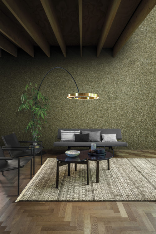 Living room in neutral colours. Wooden floor with herringbone pattern. On the walls we see Omexco's Moonstone wallcoverings, crafted from natural mica. The design is plain, yet shimmering, since mica is a semi-precious stone with alluring lustre. The color of the wall is khaki. In this picture we also see a grey sofa with pillows, two chairs, a black coffee table and a modern floor lamp.