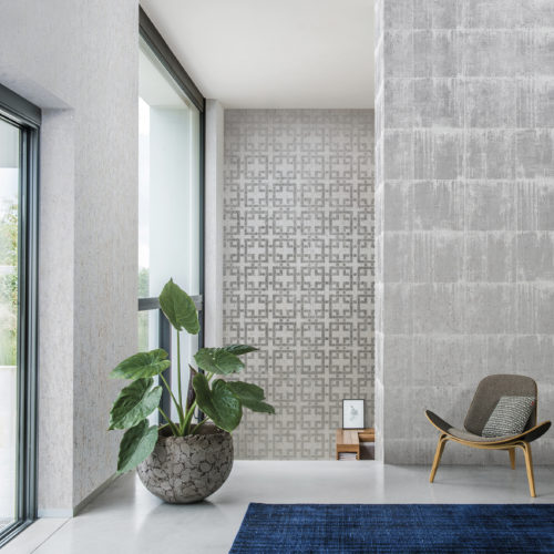 Well lit room with 3 walls of cork wallcovering. One with silver blocks, one with a silver geometrical design and one plain with golden dots. Armchair and large plant, bleu carpet.