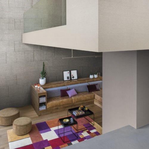 View into a colourful living room. The walls make interesting angles. On the floor a purple, orange, white, red and ocre colour blocking rug. Leather wood framed sofa.