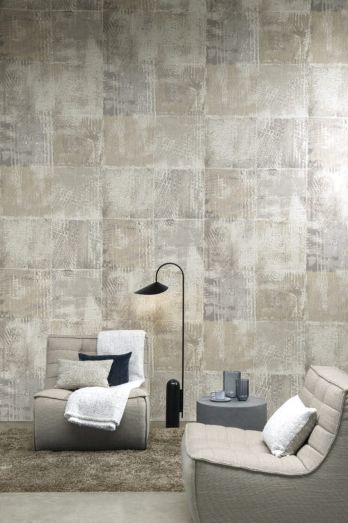 In this image we see a feature wall with organically withered squares printed on a texture of fine opalescent flakes of mica. These wallcoverings are reminiscent of graffitis faded by time. The design is called "Memoirs" and it belongs to the Moonstone collection by Omexco. Two single seater sofas are placed in front of the wall and there is a plush grey carpet on the floor. The shades in this room are neutral: beige, light grey and blue grey tones.