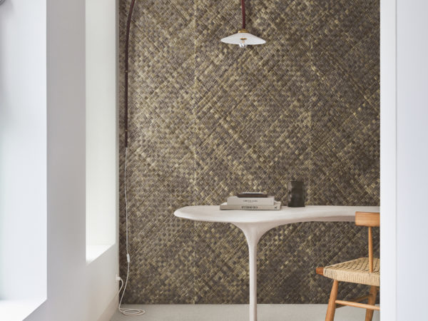 This study space features a white floor and a white wall on the left, illuminated by natural daylight streaming in through two windows. In the center of the room sits a sleek organic-shaped desk paired with a rattan chair, creating a harmonious blend of modern and natural elements. On the desk, there are several books neatly arranged. The focal point of the room is the impressive wall showcasing Omexco's handmade natural wallcoverings collection "Intuition". The wall is adorned with textured woven pandan (basket weave) in shades of brown, adding warmth and texture to the space.