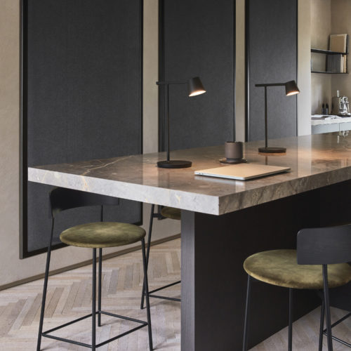 An elegant kitchen scene featuring a luxurious marble countertop adorned with sleek utensils. Two black minimalist table lamps illuminate the space, complemented by three black barstools boasting plush green velvet seating. In the background, three panels showcase Omexco's acoustic wallcovering collection, Silence, in the sophisticated color 'anthracite,' adding a touch of modernity and acoustic functionality to the chic kitchen ambiance.