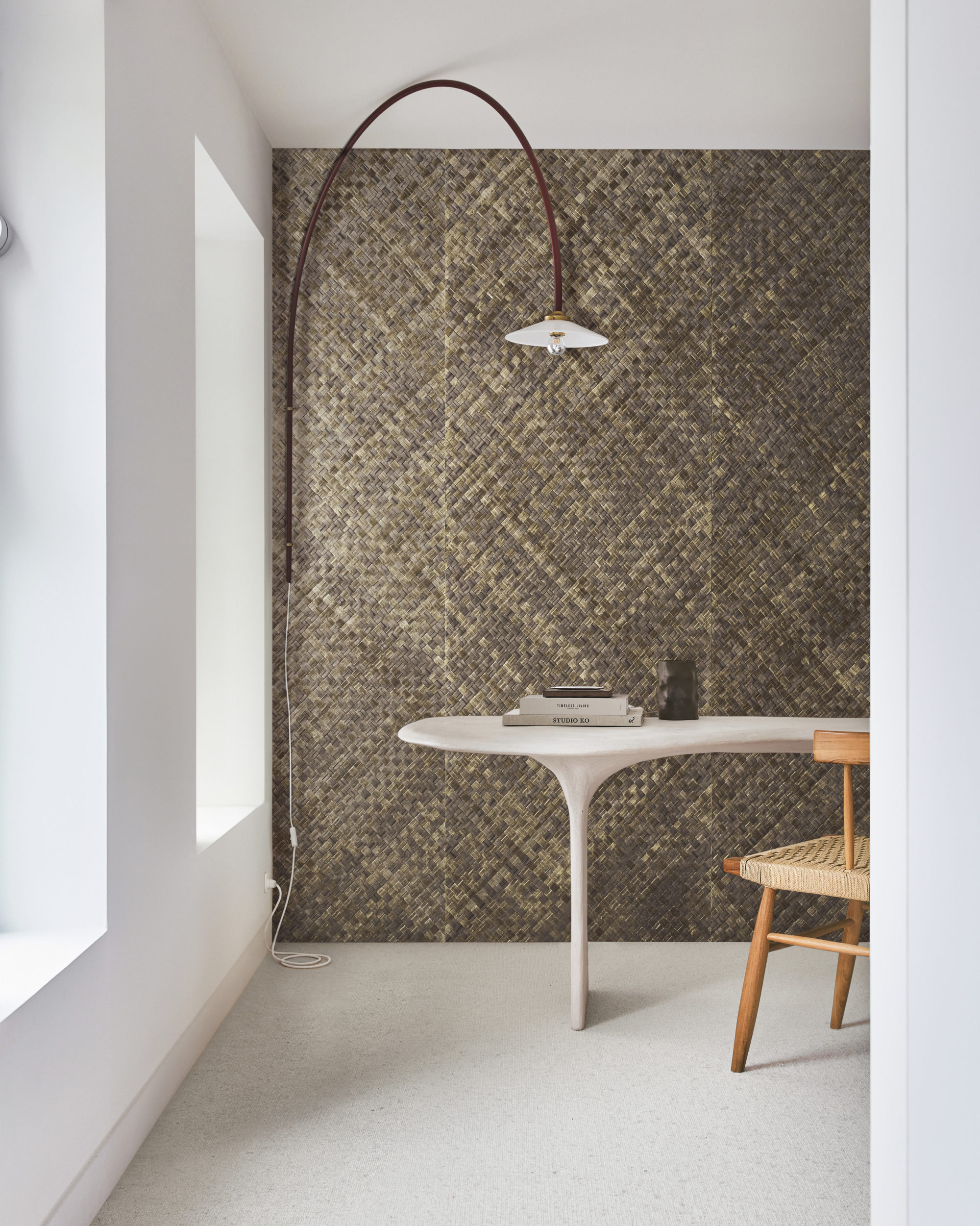 This study space features a white floor and a white wall on the left, illuminated by natural daylight streaming in through two windows. In the center of the room sits a sleek organic-shaped desk paired with a rattan chair, creating a harmonious blend of modern and natural elements. On the desk, there are several books neatly arranged. The focal point of the room is the impressive wall showcasing Omexco's handmade natural wallcoverings collection "Intuition". The wall is adorned with textured woven pandan (basket weave) in shades of brown, adding warmth and texture to the space.