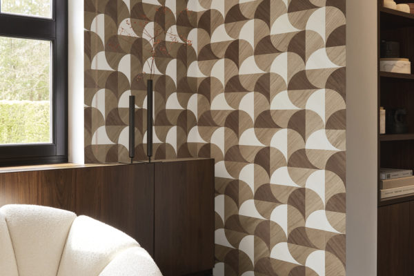 A cozy living room corner featuring a dark grey parquet floor, a white comfortable couch on the left and a window, providing natural light. On the right, built-in wall shelves display books. Adorning the wall is Omexco's marquetry collection Imperialis, showcasing geometric shapes in light brown, dark brown, and white, inspired by the graceful shape of lotus leaves.