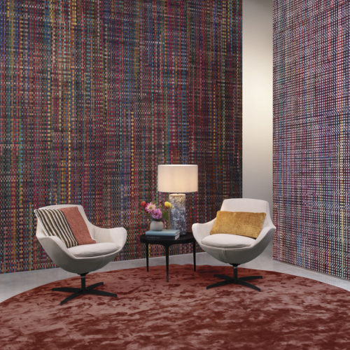 From the handweavers loom to your living room. On the walls of this impressive living room we see the "Extravaganza" wallcovering design from Omexco's Atelier collection. Colorful recycled sari silk ribbons are handwoven with Abaca. The cosy nook is also decorated with a round burgundy carpet, side table with tamble lamp, two white lounge chairs with colorful pillows and a small bouquet of fresh cut flowers.
