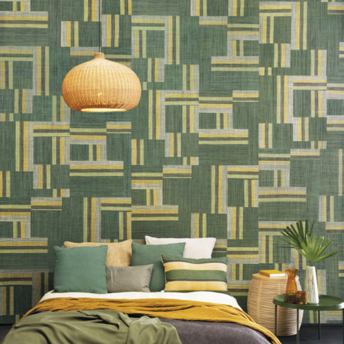 Authentic craftmanship embodies the bold Atelier collection designed by the creative minds of Omexco. This image shows an impressive green patchwork of handwoven raffia. The perfect statement wall in this tropical bedroom with ochre colour accents, a green metal bedside table, exotic plant leaf and cosy bed pillows.