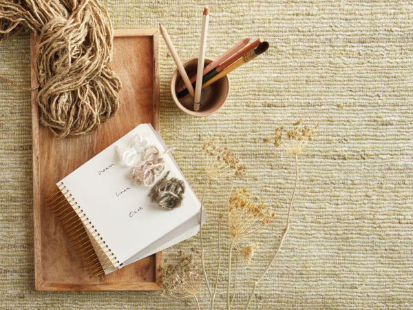 Closeup on a wooden rectangular tray on which a notebook and chunky textile yarns are posed. In the picture you can also see some dried flowers as well as a ceramic cup holding pencils. All object are posed on a large piece of chunky weave wallcoverings by Omexco. This straw-coloured product is the result of expert craftsmanship and goes straight from the handweavers loom to your interior.