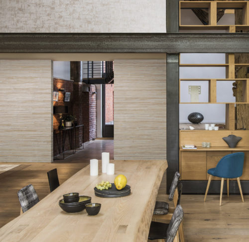Interesting dining room with iron beams, wooden table, open to the roof. In the back a hallway with brick wall. Turquoise chair.