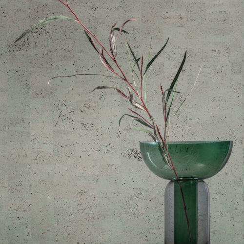 Closeup of a green transparent glass vase holding a plant stem, posed on a black round side table. In the background we see Omexco’s cork wallcovering collection “Casca", more specifically the design “tactility” in a blueish grey green colour. This is a faux plain wallcovering, the result of a patchwork of rectangular cork elements which creates a variety of different hues on the wall.
