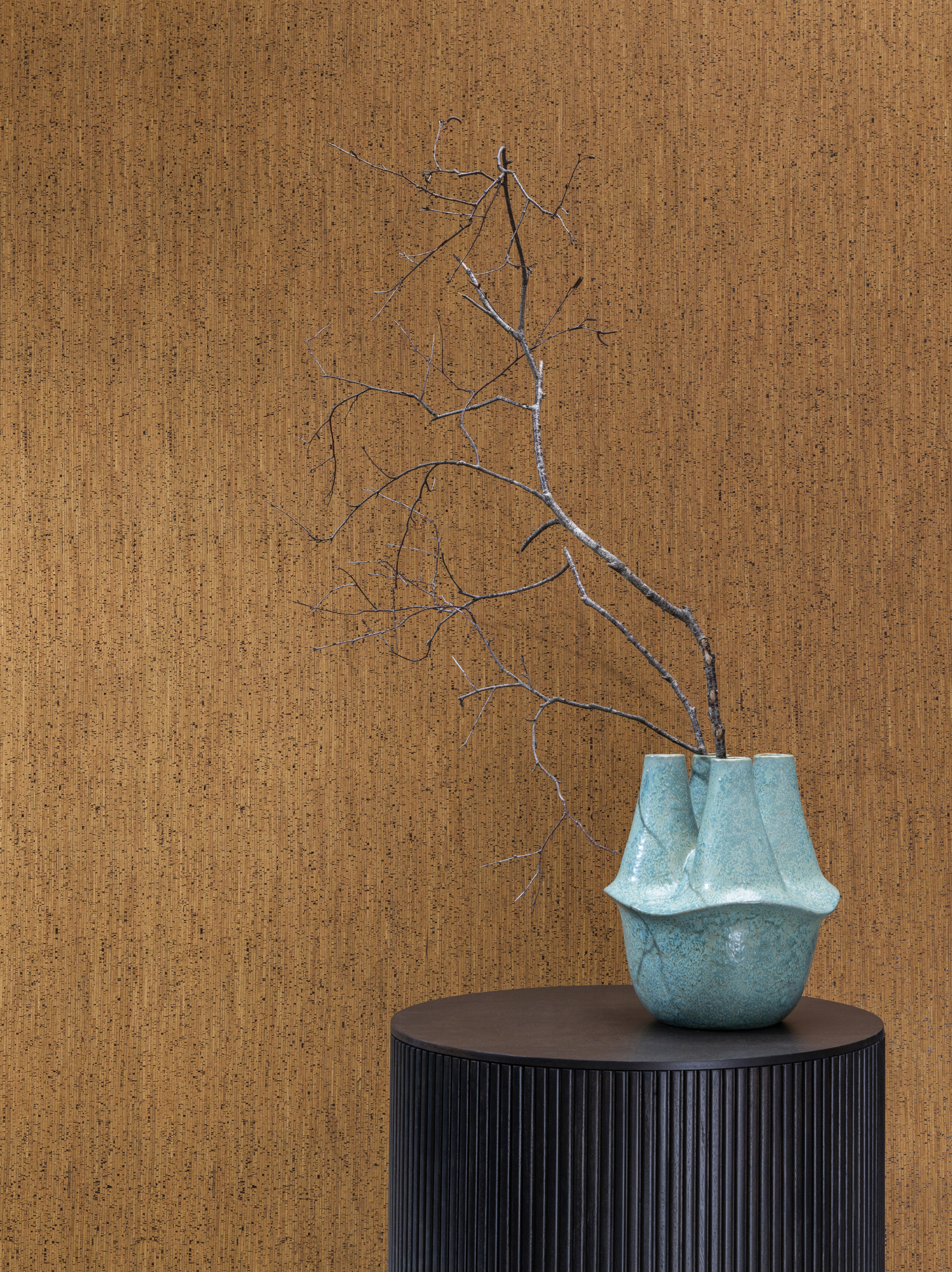 Closeup of a turquoise ceramic vase holding a small tree branch, posed on a black round wooden side table. In the background we see Omexco’s cork wallcovering collection “Casca", more specifically the design “enhance” in a caramel brown shade. This natural, handmade wallcovering is the result of many thin layers of cork that are stacked on top of each other and pressed. They are then sliced into very fine sheets. The visual result is as if the stripes are printed.